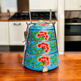 A hand-painted 3-tier steel tiffin box in blue with vibrant floral designs, displayed on a kitchen counter.