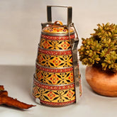 A hand-painted 4-tier steel tiffin box in gold color with intricate patterns, displayed against a neutral background with a plant to the side.