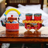 A hand-painted aluminum red tea kettle with vibrant red and yellow designs, accompanied by matching hand-painted glasses on a wooden trolley.