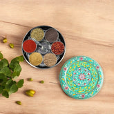A round, green hand-painted spice box with seven compartments, displayed alongside a small matching spoon and a decorative element on a wooden surface.